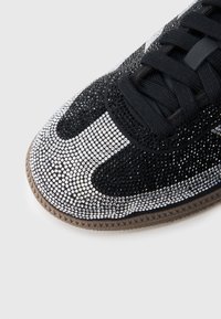 Černá teniska s třpytivým designem zdobeným černými a stříbrnými kamínky, texturovaným svrškem a gumovou podrážkou. Šňůrky jsou černé.