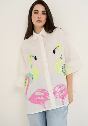 Frau trägt ein lockeres weißes Hemd mit bunten Pailletten-Flamingo-Designs und hellblaue Jeans vor einem schlichten Hintergrund.