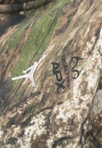 Nærbillede af camouflage-stof med et hvidt Jumpman-logo og sort "RXL" og "Legacy" tekst broderet på overfladen.