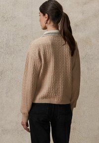 Beige kabelgebreide trui met een geribde zoom en een contrasterende kraag met zwarte strepen, gecombineerd met donkerdenim jeans.
