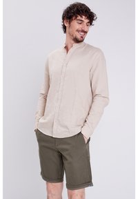 Ljusbeige långärmad skjorta med knappar och ståkrage, kombinerad med mörkgrön texturerad shorts, både avslappnad och åtsittande.