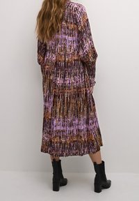 Violetti ja ruskea rypytetty maksi-mekko, jossa on eloisa abstrakti kuvio, pitkät hihat ja sulava siluetti, yhdistetty mustiin nilkkabootseihin.
