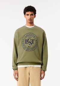 Felpa verde oliva con logo circolare blu navy "LACOSTE PARIS FRENCH SPORTSWEAR" e coccodrillo ricamato. Tessuto morbido, design a girocollo.