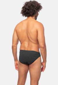 Schwarze Schwimmbrieftaschen mit gerippter Textur, niedrigem Bund und moderater Bedeckung, von hinten an einem Modell vor einem weißen Hintergrund betrachtet.