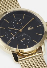 Montre à bracelet en maille dorée avec un cadran noir. Les caractéristiques incluent les heures, les minutes et des sous-cadrans pour le jour et la date, avec des accents de marqueurs dorés et un logo.
