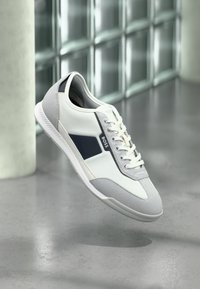 Biało-szary sneaker z sznurowadłami, czarnym bocznym paskiem i logo "BOSS", unoszący się nad szarą podłogą w pobliżu ściany z bloczków szklanych i kolumny.