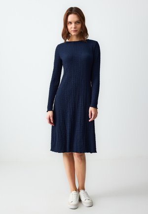 Abito blu navy a maniche lunghe con un motivo testurizzato e arricciato. Silhouette a trapezio con scollatura rotonda e lunghezza al ginocchio, abbinato a sneakers bianche.