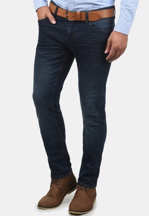Dark blue denim jeans with a slim fit, paired with a brown leather belt. A light blue long-sleeve shirt and brown ankle boots complete the look.
