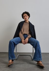 Maglione knitted marrone, cappotto blu navy, jeans svasati blu e scarpe slip-on marroni. Modello seduto su una sedia metallica contro uno sfondo semplice.