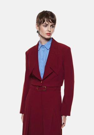 Blazer su misura bordeaux con colletto affilato, abbinato a una camicia button-up a righe blu chiaro. Include una cintura coordinata e una silhouette strutturata.