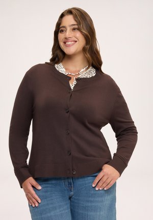 Lächelnde Frau, die einen dunkelbraunen, geknöpften Cardigan über einer gemusterten Bluse und blauen Jeans trägt, steht mit den Händen in den Hüften.
