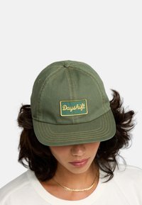 Casquette en coton verte avec une couronne arrondie, un détail de couture, et un patch rectangulaire jaune estampillé avec "Dayshift" en écriture cursive.