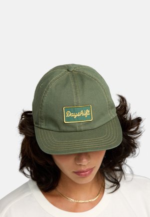 Groene katoenen pet met een ronde kroon, gestikte details en een rechthoekige gele patch die is geperst met "Dayshift" in cursief.