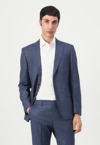 Vyriškas mėlynos spalvos tekstūruotas švarkas, pritaikytas, vienos krūtinės, su įpjovomis ir dviem priekinėmis kišenėmis, derinamas su balta marškinėlių palaidine.