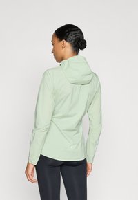 Chaqueta con capucha de color verde claro, hecha de un material ligero y suave. Presenta mangas largas y detalles reflectantes. La vista trasera muestra un ajuste relajado.