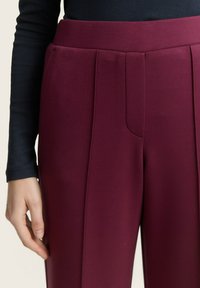 Bourgondische broek met een gladde textuur, voorzien van een hoge taille en voorkeursplooien. Het kledingstuk is gecombineerd met een donkere lange mouwtop.