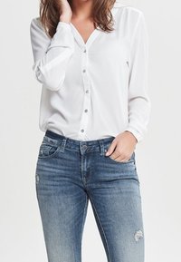 Blusa bianca a maniche lunghe con bottoni frontali, abbinata a jeans in denim chiaro e strappati con un design aderente e tasche frontali.