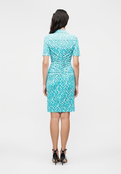 Γυναίκα με μακριά σκουρόχρωμα μαλλιά που φοράει ένα γαλάζιο και λευκό φόρεμα μέχρι το γόνατο με σχέδια και μαύρα σανδάλια με ψηλά τακούνια, στέκεται με την πλάτη προς την κάμερα.