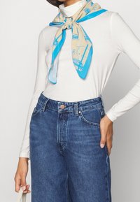 Beige Seidenschal mit blauen Akzenten, der ein grafisches Muster aufweist. Kombiniert mit einem weißen Langarmshirt und hochgeschnittenen blauen Jeans.