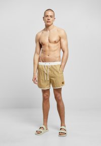 Beige badebukser med hvit trim, elastisk midje og snor. Brukes sammen med lyse, slip-on sandaler. Glatt stoff, casual passform.