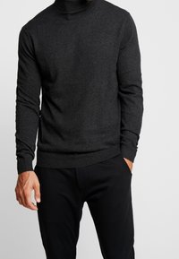 Pull à col roulé gris foncé en tissu doux, avec des manches longues et un ourlet inférieur côtelé, associé à un pantalon noir.