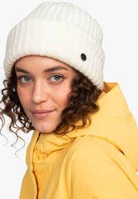 Vit ribbad stickad mössa med en svart accentknapp. Modellen bär en gul jacka med huva, delvis synlig tillsammans med mössan.