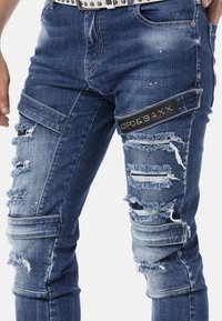 Blaue Denim-Jeans mit distressierten Patches, ausgefransten Kanten und einem schwarzen Markenlabel. Mit Details an Knien und Oberschenkeln sowie Reißverschlussakzenten.