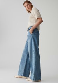 Vrouw in een witte fuzzy korte mouw trui, lichtblauwe wijde jeans met haar handen in de zakken, en witte puntige schoenen, staande op een grijze achtergrond.
