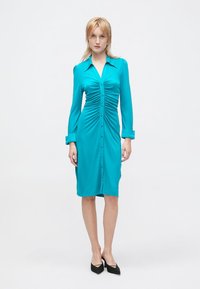 Abito button-up in teal con maniche lunghe, caratterizzato da arricciature frontali e una vestibilità su misura. Tessuto liscio con una leggera lucentezza, abbinato a scarpe nere.