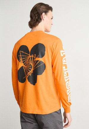 Dlouhý rukáv oranžového trička s velkým černým květinovým grafickým designem na zadní straně a bílým textem na pravém rukávu. Bavlněný materiál.