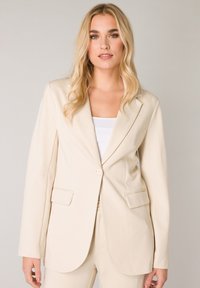 Beige blazer met twee voorzakken, enkele knoopsluiting, getailleerde revers en een soepele textuur, gecombineerd met een witte top.