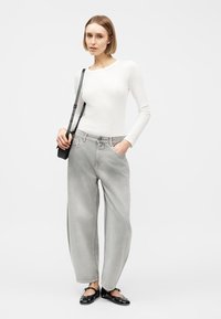 Γυναίκα που φοράει λευκό ριμπ πλεκτό τοπ με μακριά μανίκια, φαρδιά ανοιχτό γκρι τζιν, μαύρη τσάντα χιαστί και μαύρες μπαλαρίνες Mary Jane με λουράκια στον αστράγαλο.