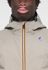 Veste K-Way beige clair avec fermeture éclair ornée de rayures rouges, jaunes et bleues, col ajustable et écusson logo sur le côté supérieur gauche.