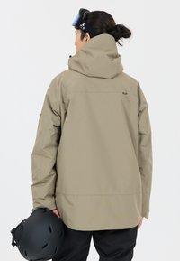 Waterdichte beige skijas met capuchon, voorzien van een relaxed fit, verstelbare manchetten en een zwarte helm in de hand.