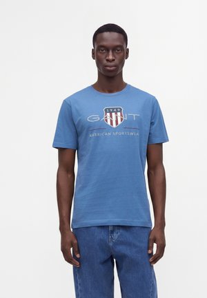 Jeune homme portant un T-shirt bleu GANT avec logo et texte, debout devant un fond blanc uni.