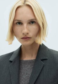 Cheveux blonds avec des vagues douces, portant un blazer gris foncé sur un pull gris texturé. Peau claire avec des taches de rousseur, fond neutre.
