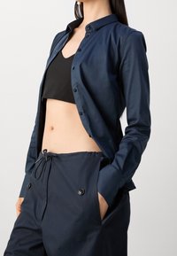 Marinblå skjorta med knappar och en mjuk textur, bärs över en svart crop top; parat med matchande marinbl å dragkedjebyxor med sidfickor.