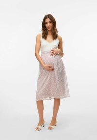 Donna incinta in un top leggero e una gonna avvolgente rosa a fantasia, che tiene delicatamente in braccio il suo pancione, sorridendo su uno sfondo semplice.