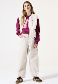 Cremefarbene Fake-Fur-Weste, burgunderfarbenes Sweatshirt mit weißer Zahl, beigen weit geschnittene Hosen und braune Sneakers mit Leopardenmuster-Akzenten.