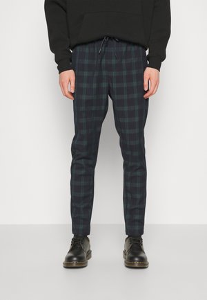 Uomo con pantaloni a quadri neri e verde scuro e stivali neri con lacci su sfondo bianco.