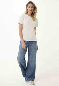 Bílý bavlněný tričko s texturovaným logem na přední straně, v kombinaci s širokými modrými džíny. Oblečení doplňují světlé boty.