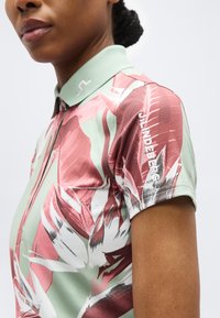 Γυναίκα που φοράει κοντομάνικη μπλούζα με γιακά, με αφηρημένα λουλουδένια σχέδια σε ροζ, λευκό και πράσινο και το κείμενο «J.LINDEBERG» στο μανίκι.