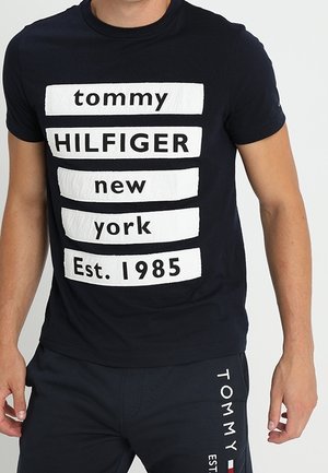 Homme portant un T-shirt noir Tommy Hilfiger avec des blocs de texte blancs indiquant "tommy HILFIGER new york Est. 1985" et un short noir assorti.