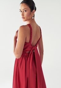 Sieviete ar tumšiem matiem, kas sasisti bulciņā, valkā sarkanu bezpiedurkņu kleitu ar savilktiem siksniņām un lielu loku aizmugurē, skatoties pāri plecam.