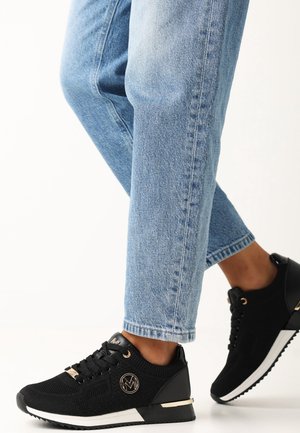 Personne portant un jean bleu clair et des baskets de sport noires avec semelles blanches, des accents métalliques et un logo circulaire sur le côté.