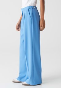 Hoge taille, wijdvallende broek van lichtblauwe stof, met plooien en een knoopsluiting aan de voorkant. Soepele textuur met een getailleerde pasvorm.