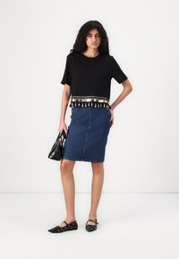 Camiseta negra corta con ribete dorado y borlas, combinada con una falda de mezclilla ajustada con botones laterales y un corte hasta la rodilla. Zapatos planos negros y bolso.