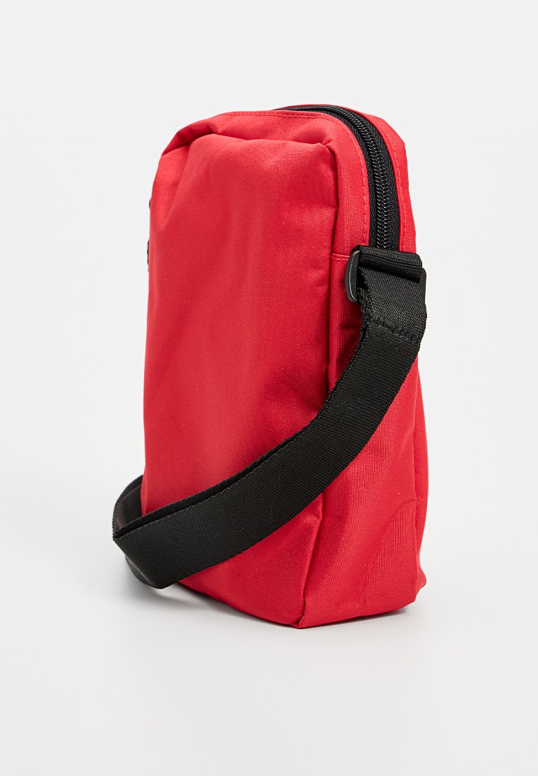 Bolso cruzado de tela roja con cierre de cremallera, forma rectangular y correa ajustable negra. Presenta una textura suave y un diseño duradero.