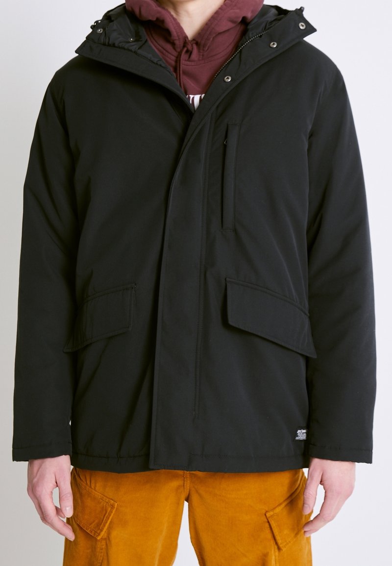 Chaqueta aislante negra con capucha, con cierre delantero, dos grandes bolsillos frontales y un bolsillo en el pecho. Textura suave, diseño minimalista.