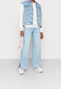 Veste en denim avec des détails usés, sweat-shirt tie-dye, jean large bleu clair avec des fentes sur les côtés, et un petit sac à main rose.